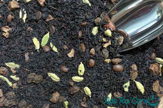 Mélange de thé noir en vrac avec grains de café et gousses de cardamome, accompagné d’une pelle à thé métallique.